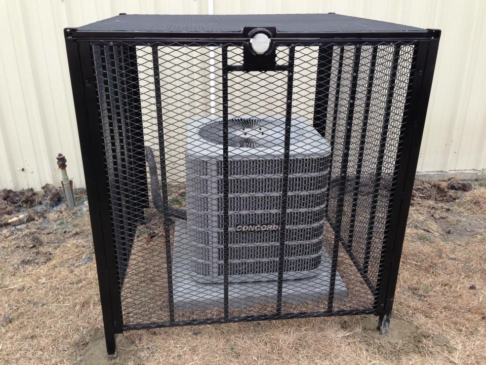 Air conditioning unit secured within a metal cage, protecting it from potential damage. The structure is placed on grass near a building exterior.