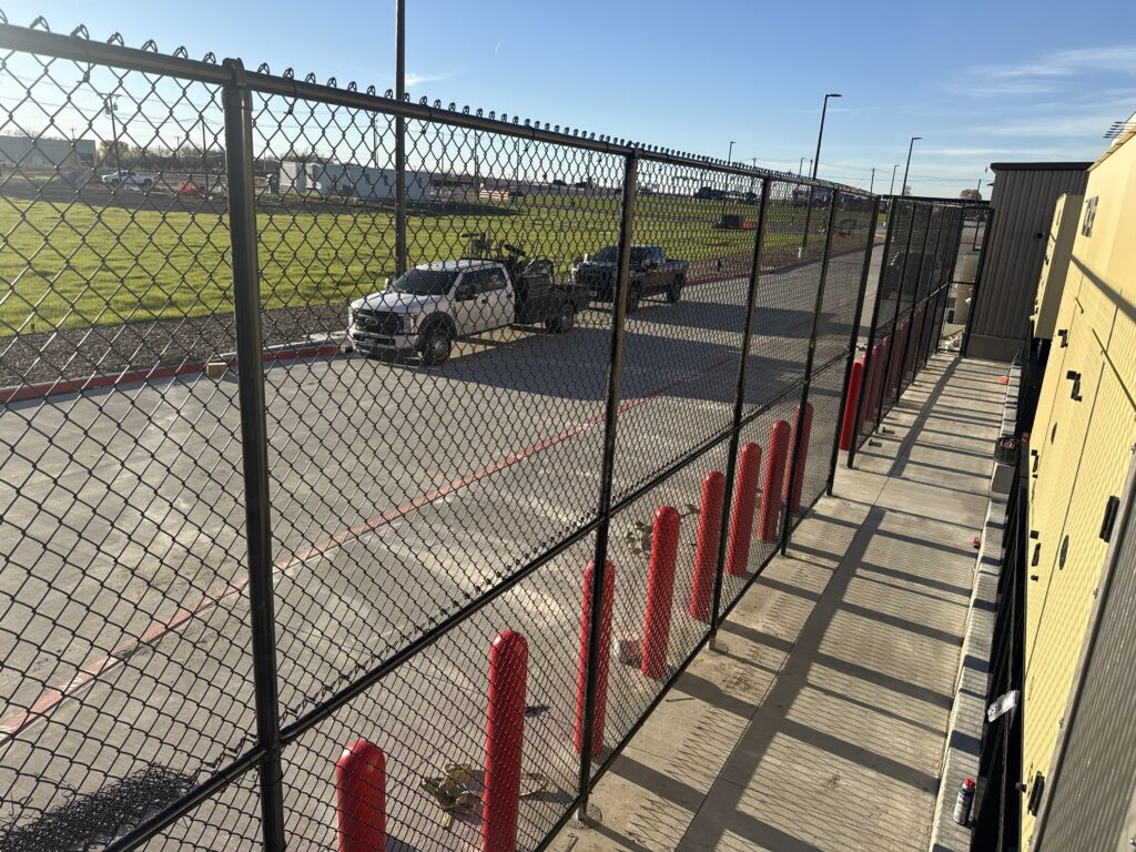 A fenced area showing two vehicles on a pavement beside red safety bollards and industrial structures, highlighting an outdoor service or loading zone.