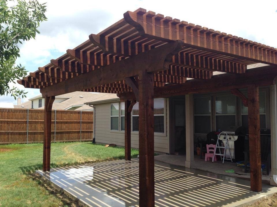 Wooden pergola with a slatted roof in a backyard, providing shade over a patio area. The space is surrounded by a wooden fence and grassy lawn.