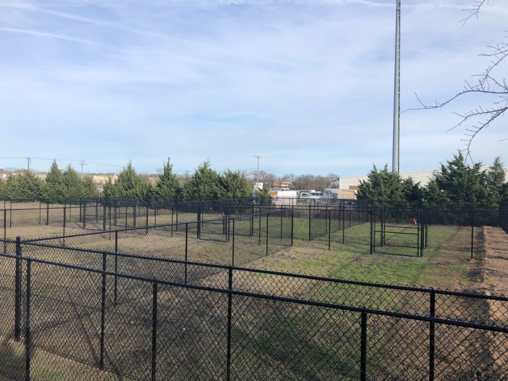 Fenced dog park with multiple enclosed grassy areas and evergreen trees, providing a space for pets to play and exercise safely.