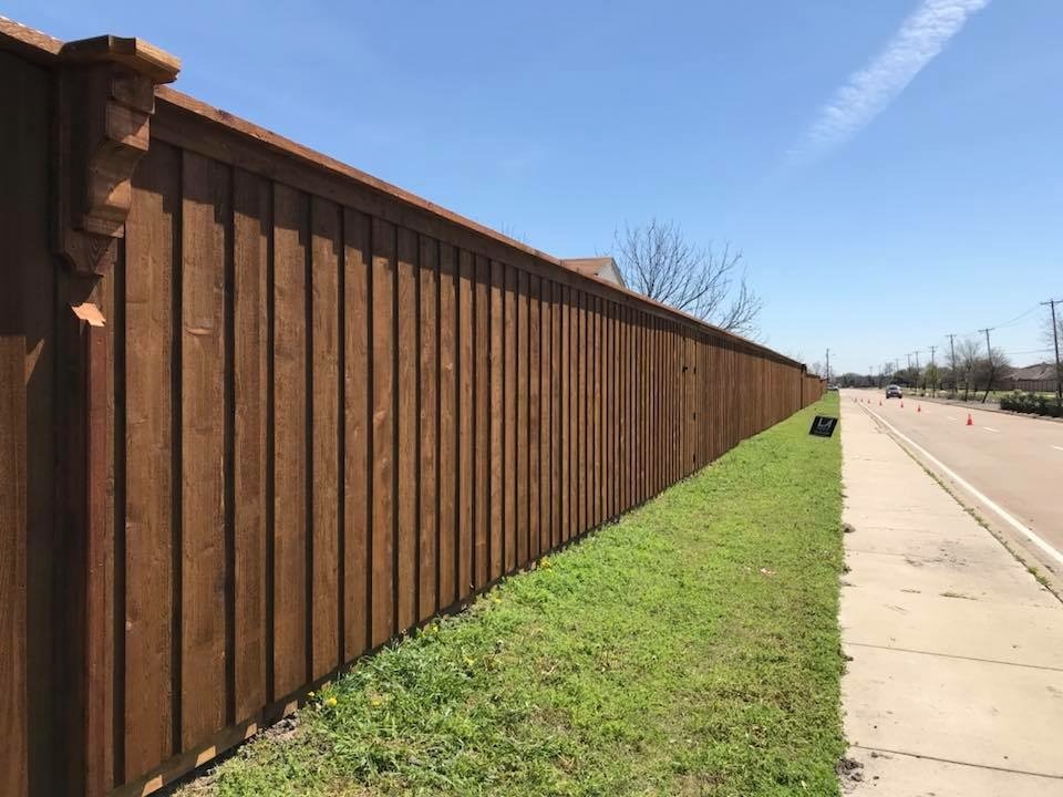 A long wooden fence runs alongside a grassy area and a paved road under a clear blue sky, indicating property boundaries and outdoor space.