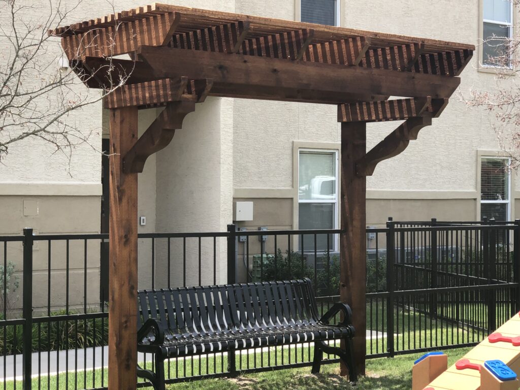 Wooden pergola over a black metal bench in a landscaped area, providing shaded seating near a fenced play space. Ideal for relaxation and outdoor gatherings.